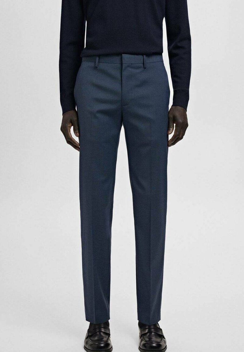 Mężczyzna ubrany w dopasowane granatowe spodnie garniturowe, czarną bluzkę z długim rękawem oraz czarne skórzane eleganckie buty, stojący na jednolitym tle.