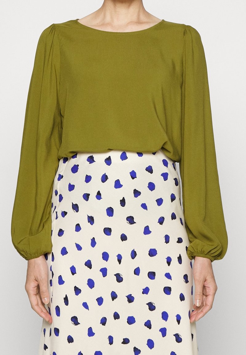 Femme portant un chemisier vert olive à manches longues rentré dans une jupe blanche avec des taches bleues et noires irrégulières.