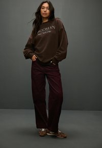 Barna kapucnis felső "BROOKLYN New York City" felirattal, burgundi kordbársony nadrággal és leopárd mintás sportcipővel párosítva. Simább textúrák, laza illeszkedés.