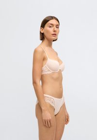 Gaiši rozā apakšveļas komplekts ar mežģīņu krūšturi ar plānām lencītēm un atbilstošām biksītēm ar mežģīņu dekoru, izceļot smalkas rakstu un tekstūru detaļas.