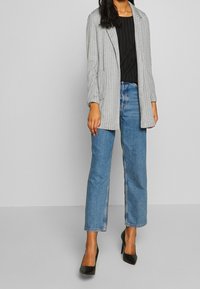 Mujer vistiendo un blazer gris de rayas, blusa negra, jeans rectos azules y zapatos de tacón negros, de pie contra un fondo liso.