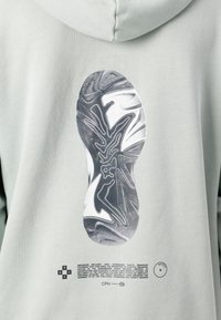 Helle grüne Kapuzenjacke mit einem schwarz-weißen Grafikdesign eines Sneaker-Sohlen und Text, mit einer glatten Textur und minimalistischem Design.