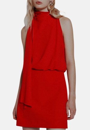 Vestito rosso senza maniche con collo alto e dettaglio in tessuto drappeggiato indossato da una donna con lunghi capelli biondi e orecchini a forma di stella.