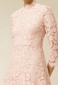Krajkované šaty v jemně růžové barvě s vysokým límcem a tříčtvrtečními rukávy, s květinovými vzory a vlnitými okraji. Elegantní a texturovaný design.