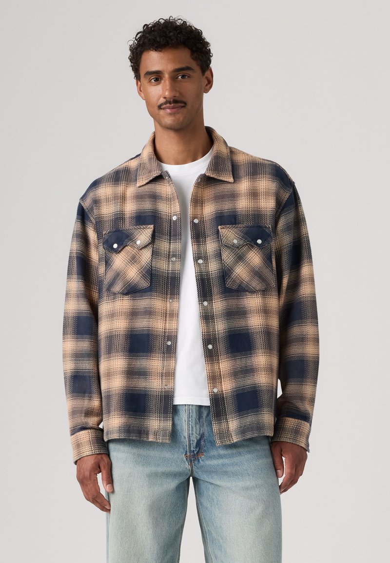 Homme portant une chemise à carreaux beige et marine boutonnée par-dessus un t-shirt blanc et un jean bleu clair sur un fond uni.
