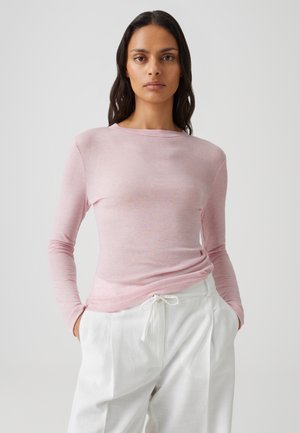 Vrouw met donker haar die een lichtroze longsleeve en witte broek met trekkoord draagt, staand met handen in de zakken tegen een effen achtergrond.