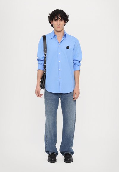 Hellblaues Button-Up-Hemd mit kleinem schwarzem Logo auf der linken Brust, kombiniert mit blauen Jeans im geraden Schnitt und schwarzen Schuhen, während eine schwarze Tasche gehalten wird.