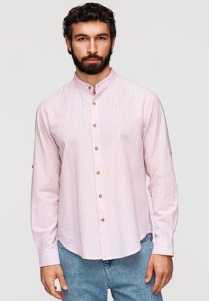 Homme avec barbe portant une chemise à col mandarin rose clair à fines rayures et un jean bleu, debout devant un fond uni.