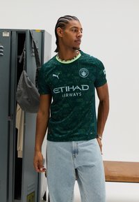 Groen voetbalshirt met geometrische patronen, korte mouwen en een ronde halslijn. Voorzien van merk- en reclametekst. Draagt lichtblauwe jeans.