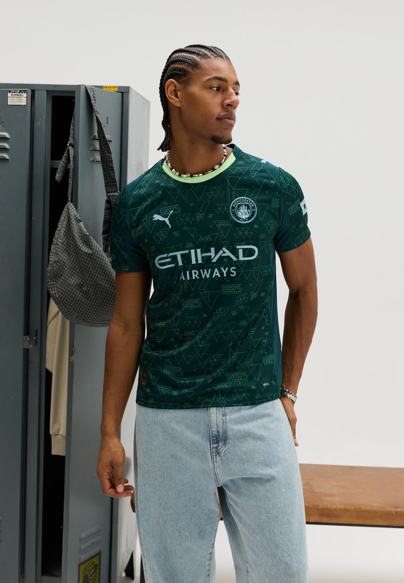 Groen voetbalshirt met geometrische patronen, korte mouwen en een ronde halslijn. Voorzien van merk- en reclametekst. Draagt lichtblauwe jeans.