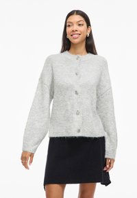 Pilkos vilnonės kardiganas su apvaliu iškirpimu, priekiniais mygtukais, nuleistais pečiais ir minkštu, tekstūruotu paviršiumi; dėvimas virš juodos sijono.