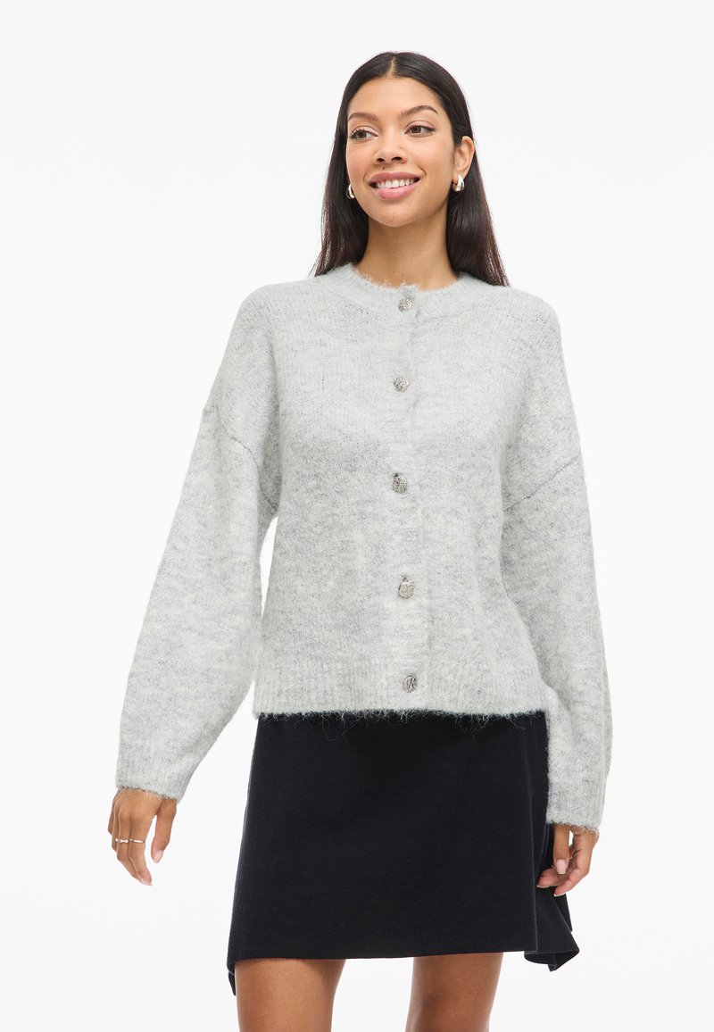 Pilkos vilnonės kardiganas su apvaliu iškirpimu, priekiniais mygtukais, nuleistais pečiais ir minkštu, tekstūruotu paviršiumi; dėvimas virš juodos sijono.