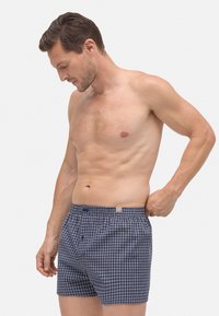 Hombre adulto ajustándose los pantalones cortos de cuadros azul y blanco, de pie sin camiseta contra un fondo blanco liso.