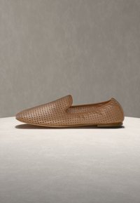 Ταν δερμάτινο loafer τύπου slip-on με διάτρητο σχέδιο και χαμηλό τακούνι, τοποθετημένο πάνω σε ανοιχτό γκρι επιφάνεια με ουδέτερο φόντο.