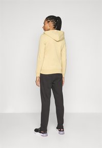 Gelbe Kapuzenjacke mit Kordelzug, gerippten Bündchen, kombiniert mit schwarzen Jogginghosen. Das Outfit beinhaltet schwarze Sneaker mit rosa und lila Akzenten.