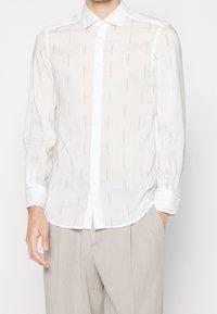 Biała koszula z długim rękawem, wykonana z lekkiej tkaniny, z subtelnym powtarzającym się wzorem. Standardowy kołnierzyk i mankiety zapinane na guziki.