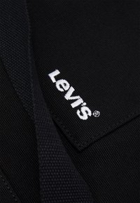 Svart tyg med en strukturerad yta som har en vit broderad "Levi's" logotagsetikett. Fokus på logotypen och tygets textur och färg.