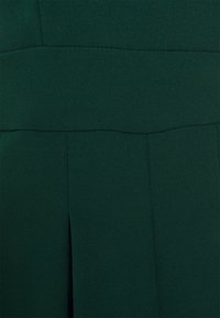 Tmavě zelené šaty s texturovanou tkaninou, které mají horizontální šev v pase a svislý rozparek na spodní části.