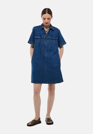 Robe chemise en denim bleu foncé avec des manches courtes, des poches sur la poitrine, un détail de lacets au niveau du décolleté et une coupe lâche et droite.