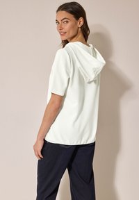 Haut à capuche blanc texturé avec des manches courtes, présentant une coupe décontractée et un tissu doux, associé à un jean foncé.