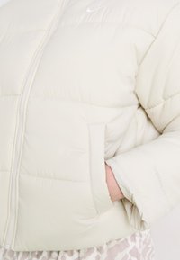 Giacca puffer bianca con design trapuntato, chiusura con zip e tasche laterali. Presenta il logo Nike e il marchio Therma-FIT sulla manica.