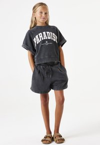 Grijze cropped sweatshirt met de tekst "PARADISE", gecombineerd met passende high-waisted shorts. De stof lijkt zacht; de schoenen zijn bruin met decoratieve accenten.