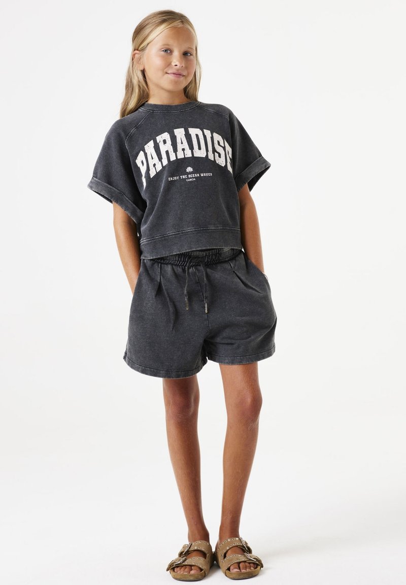 Grijze cropped sweatshirt met de tekst "PARADISE", gecombineerd met passende high-waisted shorts. De stof lijkt zacht; de schoenen zijn bruin met decoratieve accenten.