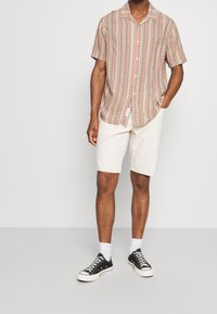 Mies, joka on pukeutunut lyhythihaiseen, nappikiinnitteiseen raitapaitaan, valkoisiin polveen ulottuviin shortseihin, valkoisiin sukkiin ja mustiin matalakärkisiin lenkkareihin, seisoo valkoista taustaa vasten.