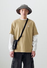 Tan kortærmet t-shirt med logo, lagdelt over en hvid langærmet skjorte. Sort taske med skulderrem, grå bredskygget hat, mørke cargo bukser.