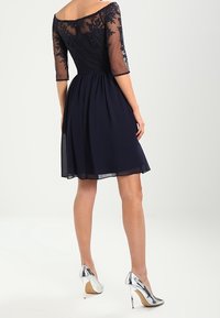 Mörkblå klänning med en figurnära bodice, genomskinliga trekvartslånga ärmar och broderade detaljer, som övergår i en plisserad kjol. Bärs med silverfärgade klackar.