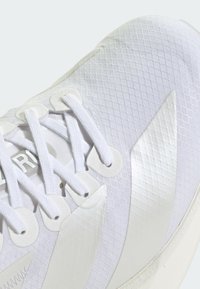 Chaussure de sport blanche avec un dessus texturé à motif en diamant, lacets plats et accents brillants sur le côté.