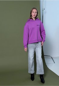 Fioletowy hoodie z przednią kieszenią i logo, zestawiony z jasnoszarymi szerokimi dżinsami oraz czarnymi botkami, na neutralnym tle.