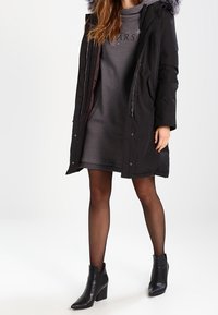 Parka negra con capucha forrada de piel, que presenta bolsillos con cremallera y cierres a presión. Llevada sobre un vestido gris, con botines negros y pantys transparentes.