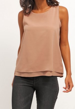 Blouse sans manches, marron clair, à couches, portée avec un jean gris foncé ajusté sur une personne debout sur fond uni.