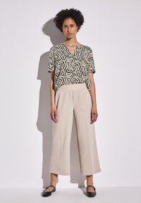 Donna con capelli ricci, che indossa una maglietta a maniche corte con motivo bianco e nero, pantaloni cropped beige a gamba larga e scarpe Mary Jane nere, in piedi contro un muro bianco.