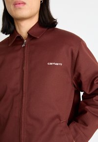 Brun zip-up jacka i slät tyg med krage och broderad logotyp på bröstet. Har synlig sömnad och en fungerande dragkedja.