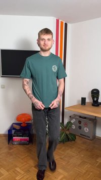 Zelena bombažna majica s belim grafičnim motivom na levi prsni strani, kombinirana s temno sivimi kavbojkami z ravnimi hlačnicami in rjavimi čevlji. Ozadje lesen pod.