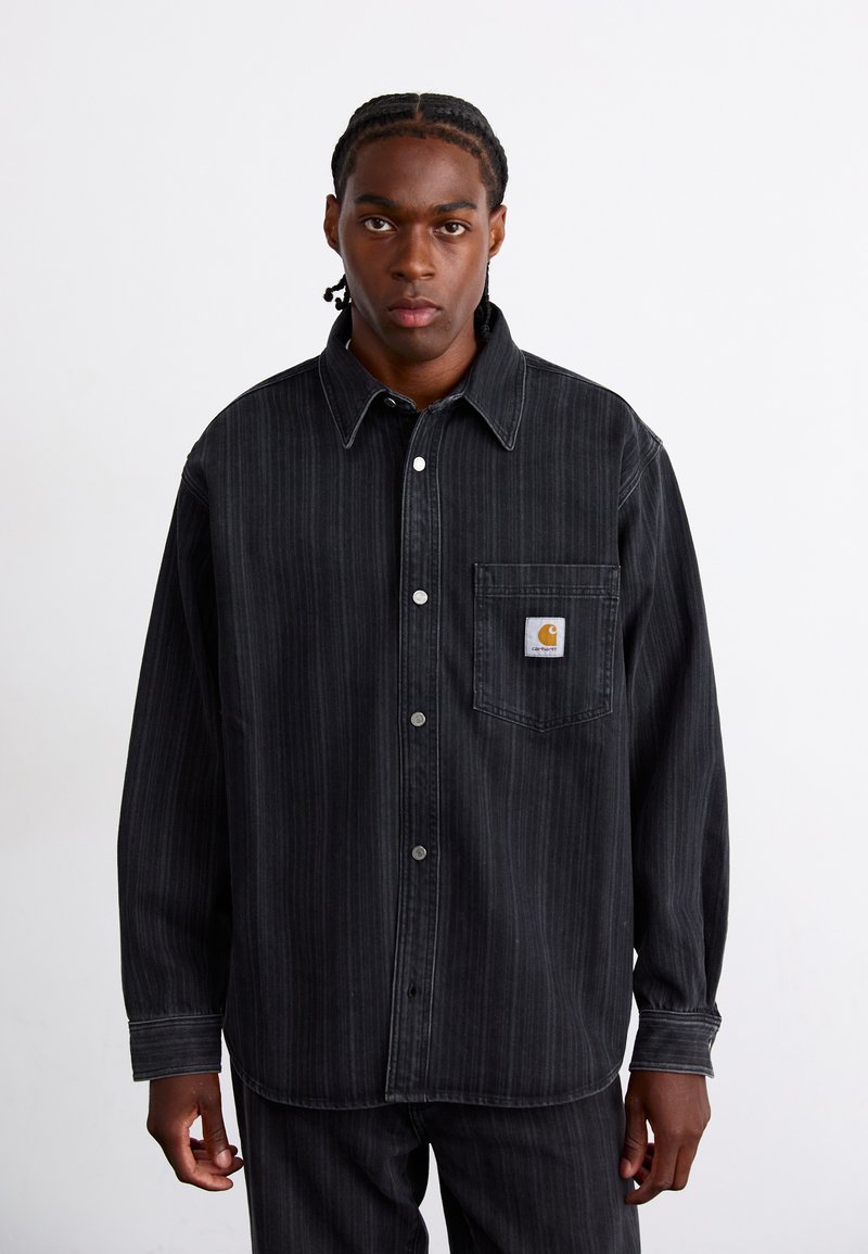 Camicia in denim nero con strisce verticali, design con bottoni, colletto e tasca frontale con patch a forma di quadrato. Indossata con pantaloni simili.