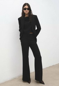 Blazer noir cintré et haut court assortis à un pantalon évasé. Accessoires : lunettes de soleil noires étroites et chaussures foncées à bout pointu.