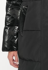 Zwarte pufferjack met glanzende afwerking. Beschikt over een getextureerde linkermouw en een zijzak. Zacht, geïsoleerd materiaal met een gevlochten patroon.