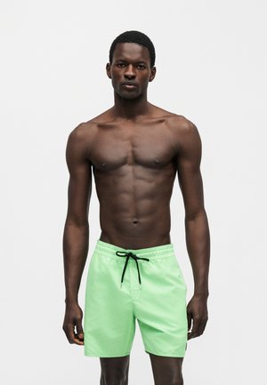 Muskulózny černoch stojaci na jednoliatom pozadí, oblečený v svetlozelených plavkách s čiernymi šnúrkami, smerujúci dopredu s neutrálnym výrazom.