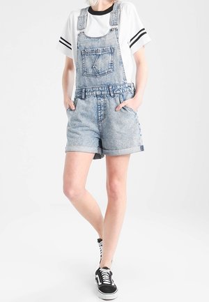 Henkilö pukeutunut haalistuneisiin sinisiin farkku shortsidezhardeihin, valkoiseen t-paitaan, jossa mustat raidat hihoissa, ja mustavalkoisiin lenkkareihin, seisoo käsin taskuissa.