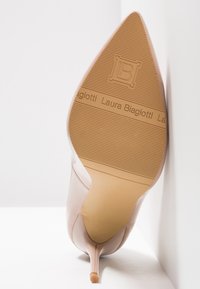 Nude högklackad sko med spetsig tå, slät ovandel och texturerad gummi-sula med varumärke och horisontella linjer.