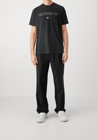 Camiseta negra de manga corta con el gráfico "HILFIGER '85" y un pequeño logotipo de bandera multicolor en el pecho, combinada con pantalones negros y zapatillas blancas.