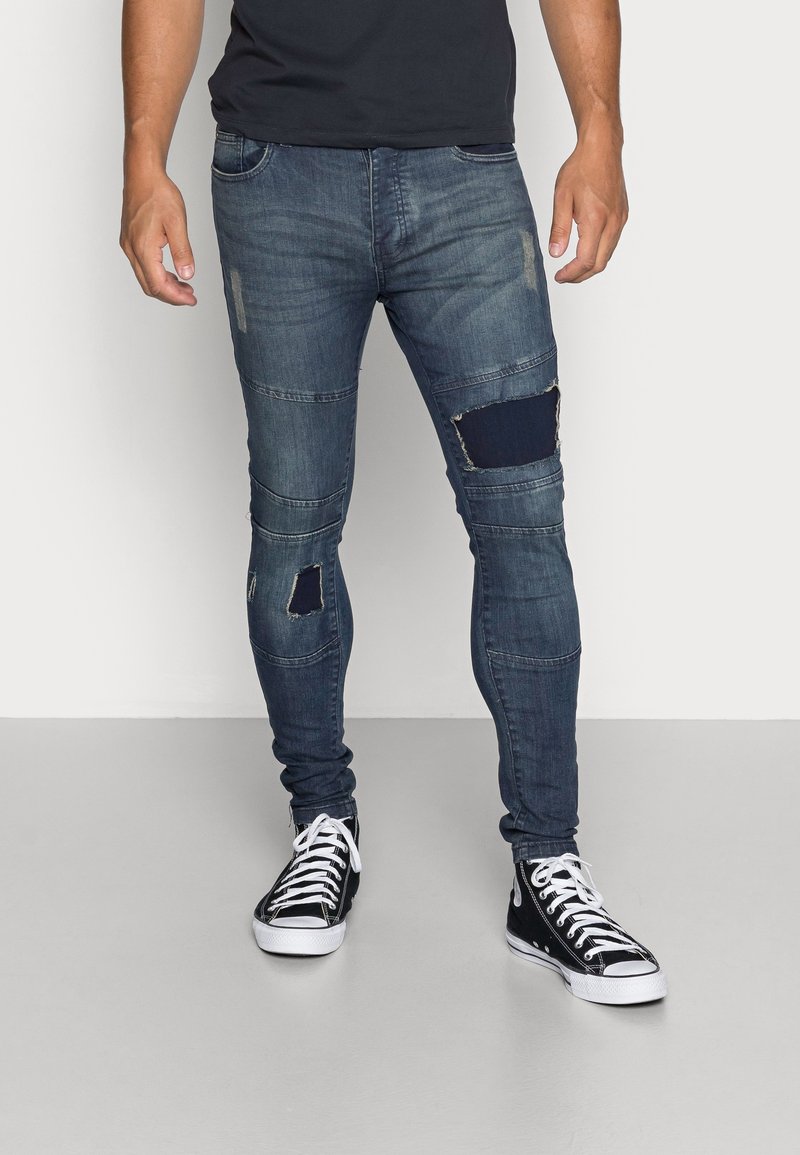 Rippade denimjeans i mörkblått med lappar, med slim fit, detaljerad sömnad och fransade kanter. I kombination med svarta högknäppta sneakers.
