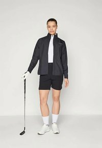 Must have golfimantli musta, millega on ühendatud mustad lühikesed püksid. Seljas valge T-särk ja valged spordijalatsid, hoides golfikeppi.