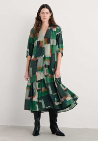 Mujer vistiendo un vestido midi de patrón abstracto verde con mangas de tres cuartos y botas negras hasta la rodilla, de pie contra un fondo liso.