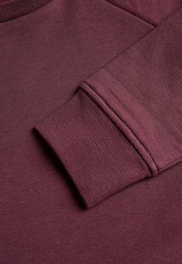 Suéter burdeos con cuello redondo y puños acanalados, fabricado en un tejido suave con una textura suave, resaltando intrincados detalles de costura.