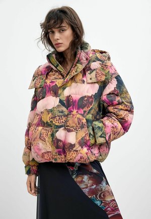 Frau trägt eine bunte, floral gemusterte Daunenjacke mit hohem Kragen und Klappentaschen, kombiniert mit einem dunklen Rock mit Blumenmotiven.