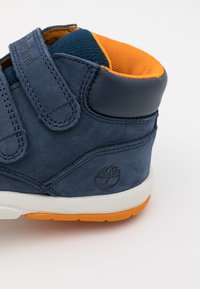 Pantofii înalți din piele bleumarin cu curele Velcro, căptușeală respirabilă din plasă portocalie, talpă intermediară albă și tălpică din cauciuc portocalie. Prezintă logo-ul mărcii.